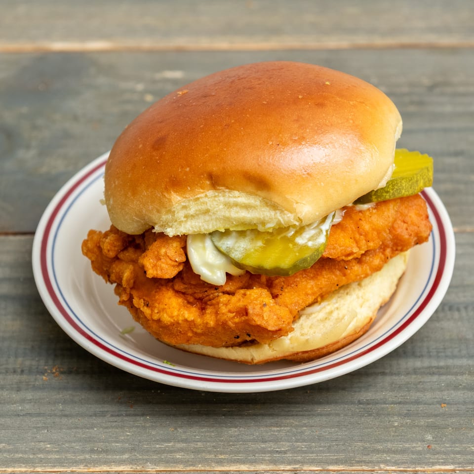 Crispy fried chicken sandwich with pickles and mayonnaise on a toasted bun, served on a plate atop a wooden table.