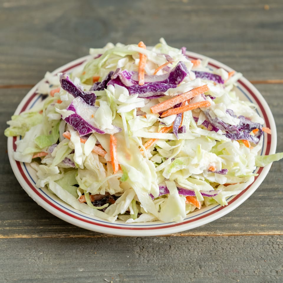 A plate of creamy coleslaw with shredded cabbage, purple cabbage, and carrots on a rustic wooden table.