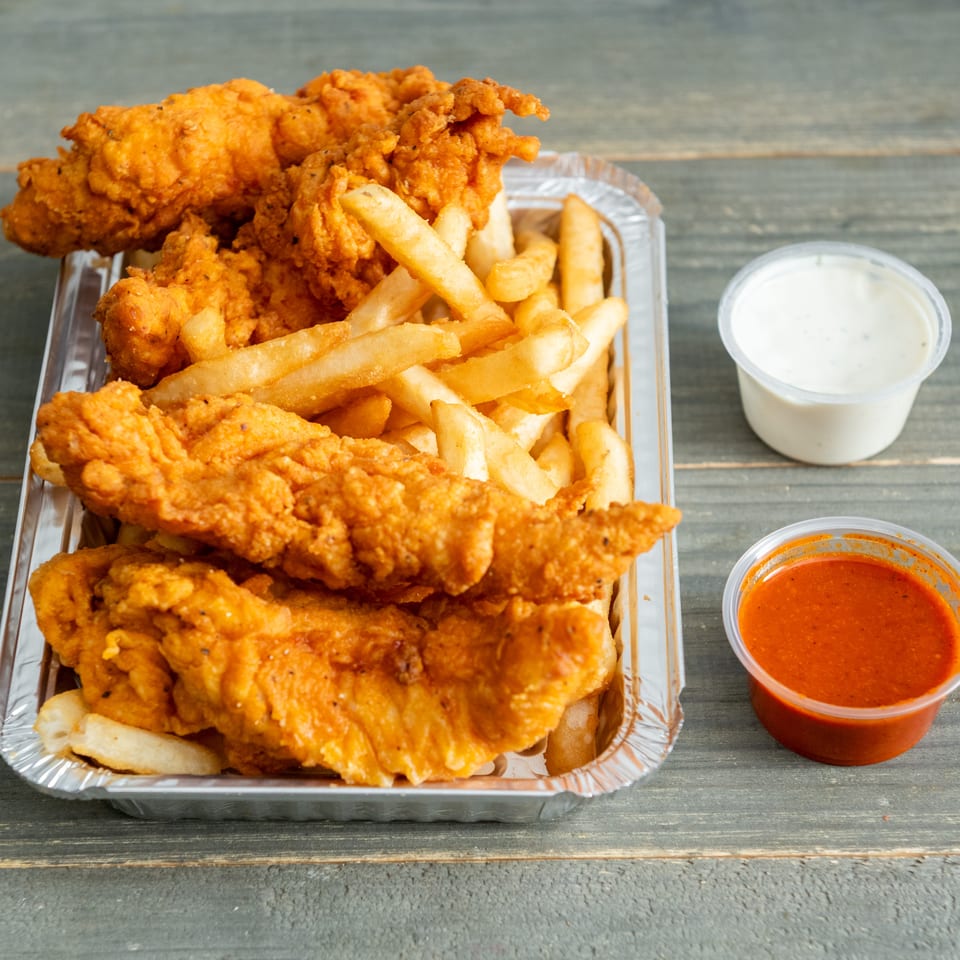 Crispy fried chicken tenders and French fries in a foil tray with white and red dipping sauces on a wooden table.